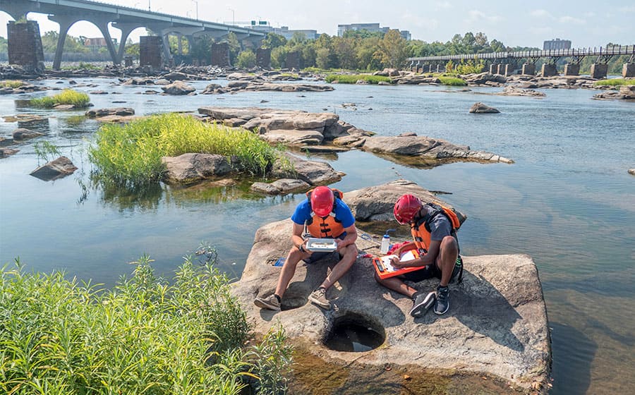 Students sit on rocks in river while taking water samples.