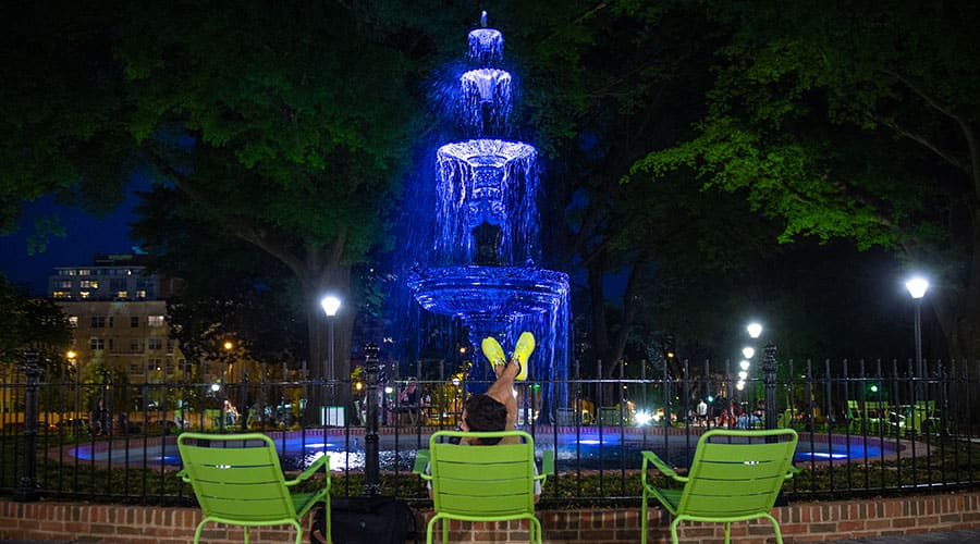 A person reclines in a chair in front of a lit-up fountain at night