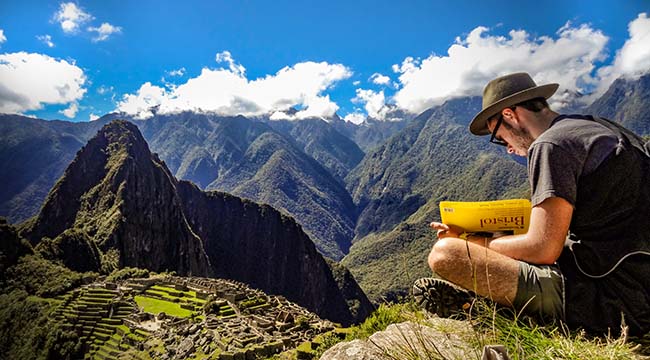 Student with sketchbook sitting on hilltop with Peruvian ruins and mountains in the distance