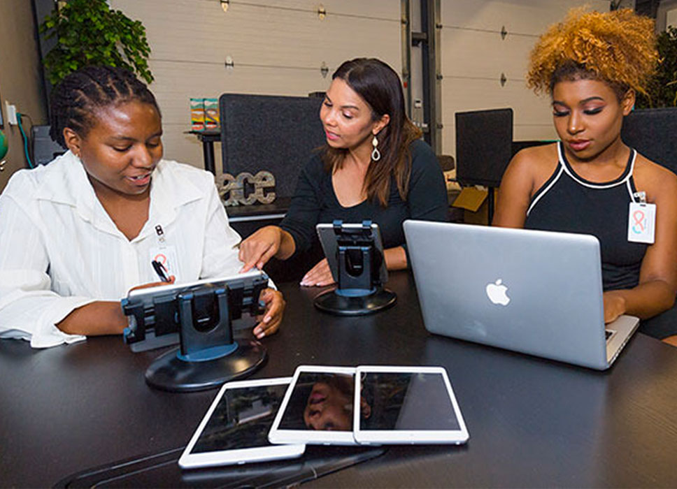 Three women, one in a lab coat, working on iPads and laptops